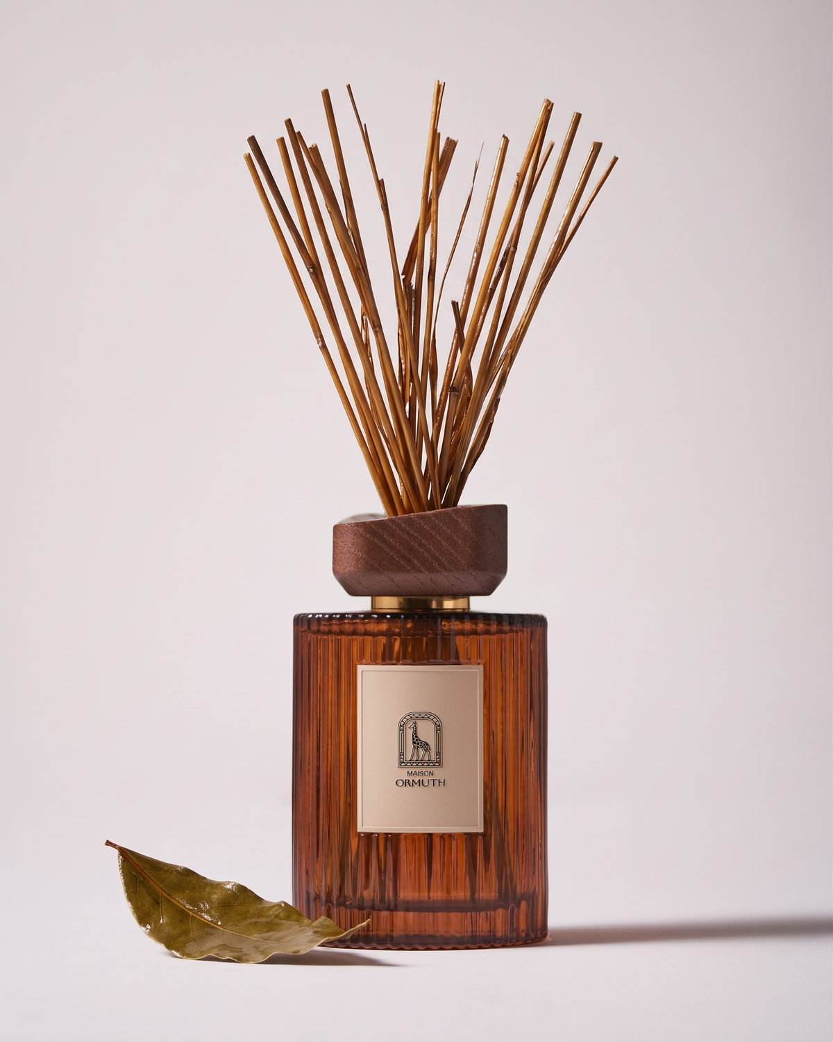 Sovereign Hay reed diffuser by Maison Ormuth: ribbed amber glass vessel with a walnut cap, reeds the color of dried hay and a single dried leaf at its base