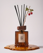 Noble Sap reed diffuser by Maison Ormuth: ribbed amber glass vessel with black reeds and a raspberry cane, standing in a pool of sap