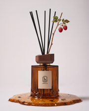 Noble Sap reed diffuser by Maison Ormuth: ribbed amber glass vessel with black reeds and a raspberry cane, standing in a pool of sap